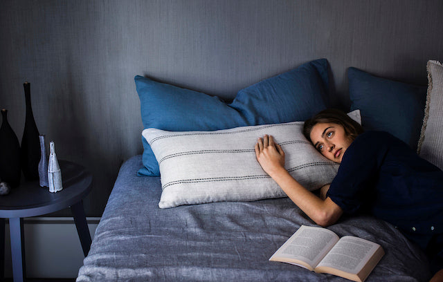 A woman lies on a bed hugging a pillow, looking thoughtful. An open book rests in front of her. The bedding and walls are in cool blue and gray tones, creating a calm, introspective scene.