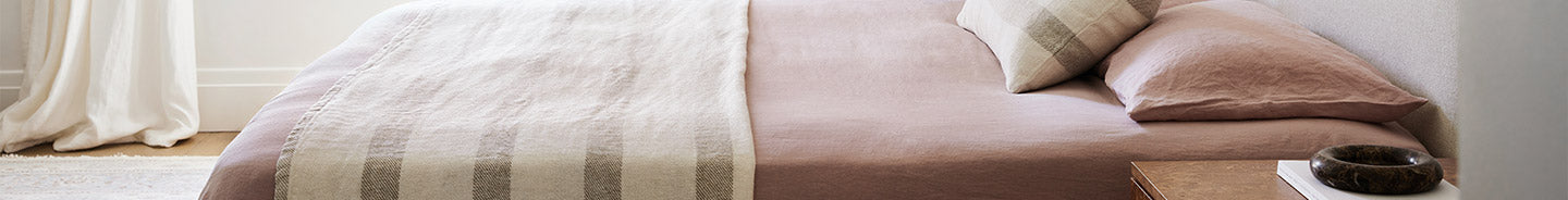 A neatly made bed with muted pink bedding, a striped beige and gray blanket, and a matching pillow. There is a small wooden nightstand with a bowl and a book beside the bed. Light curtains hang by the window.