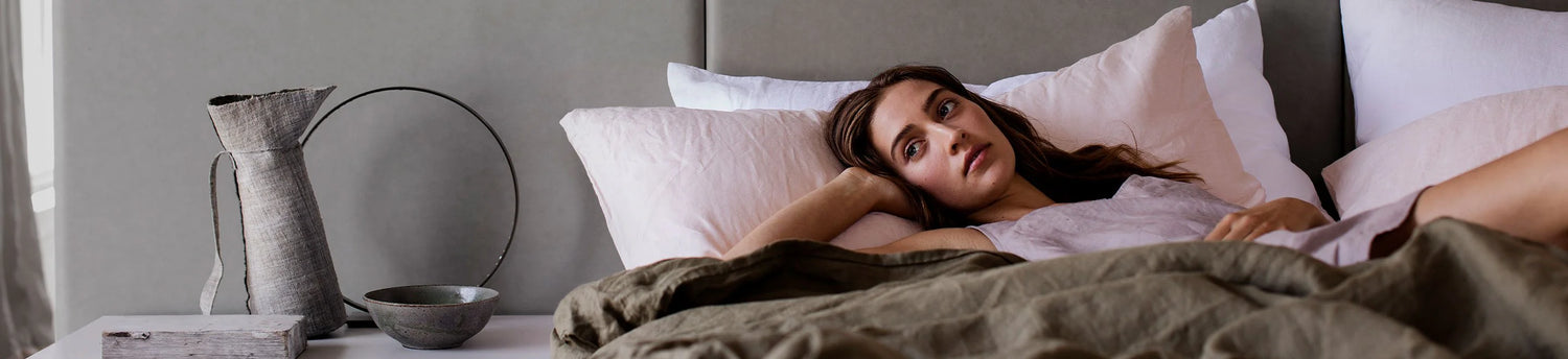 A woman lies in bed, propped up on one arm, covered with green bedding. She gazes thoughtfully ahead. A grey jug and bowl sit on the bedside table against a neutral background.