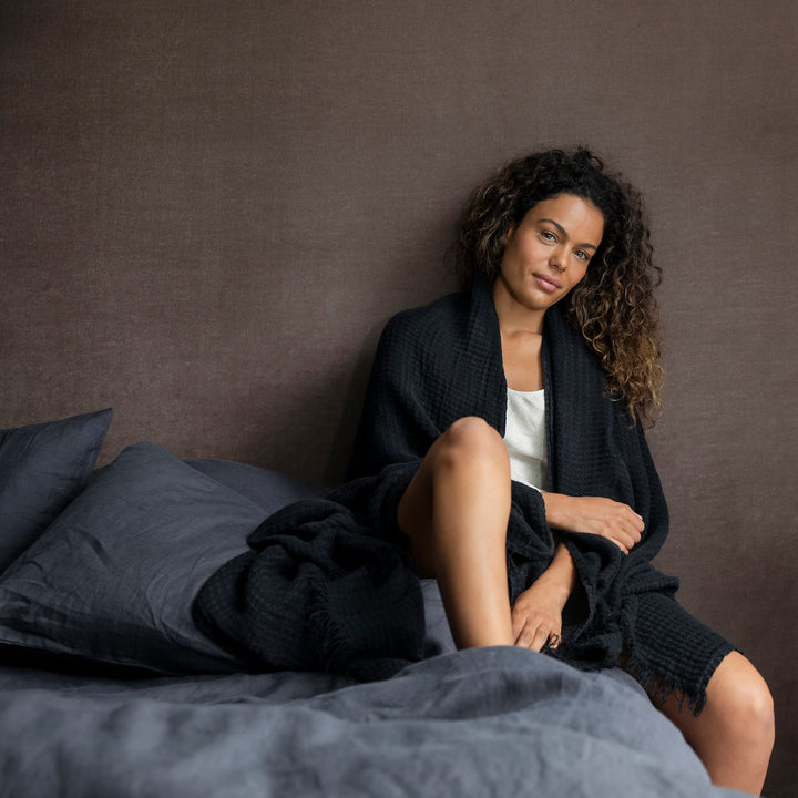 A woman is sitting on a bed dressed in Slate bedlinen, she is wearing a Black Linen Waffle Robe.