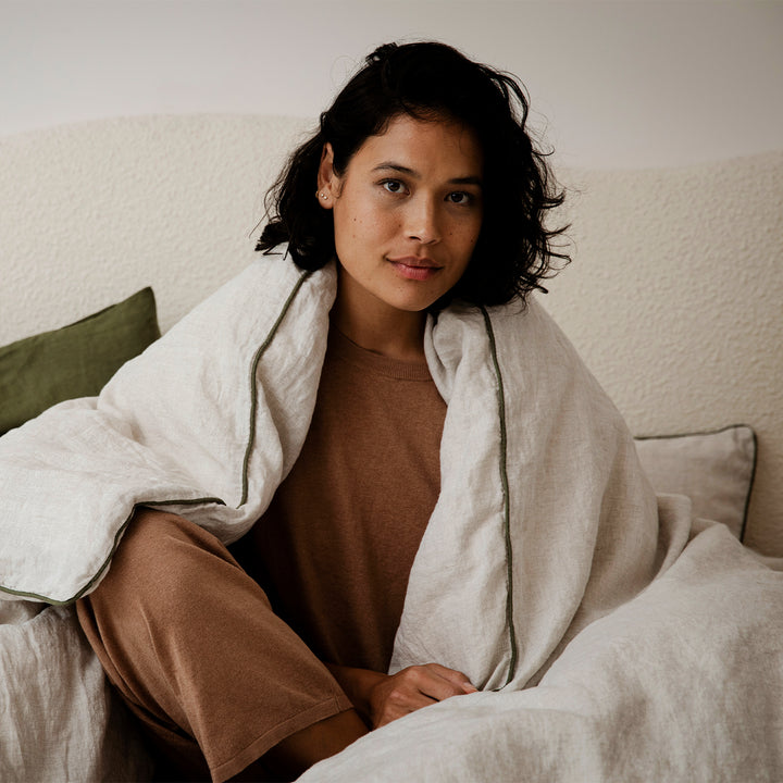 A woman sitting on a bed, with a Natural and Forest Piped linen Duvet Cover over her shoulders