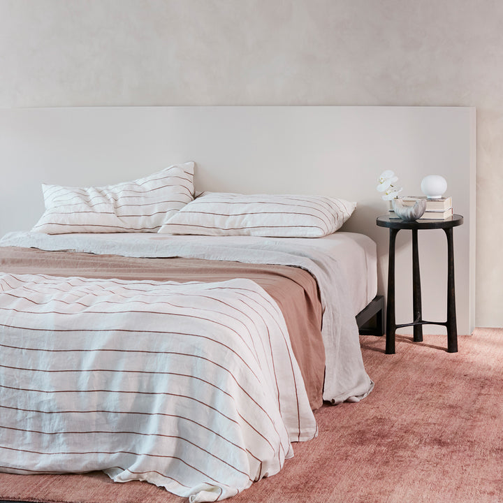 A bed dressed in Cedar Stripe, Smoke and Fawn bedlinen, styled with a dark side table.
