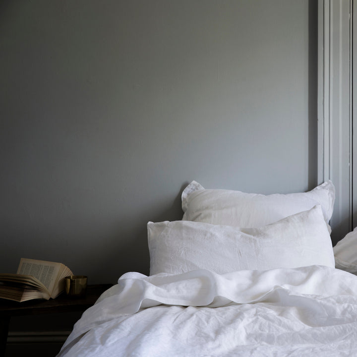 A bed dressed in White bedlinen, is styled against gray walls and a book on a side table.