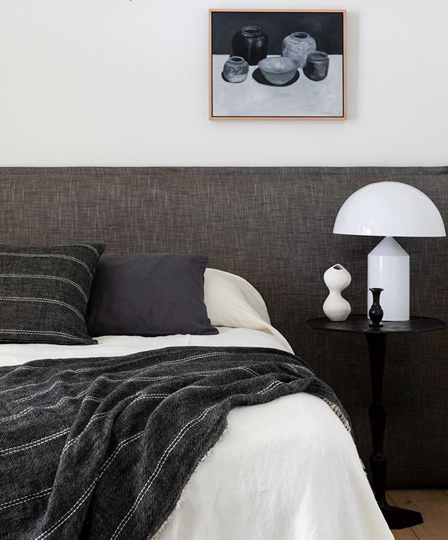 A modern bedroom features a gray upholstered headboard, striped gray bedding, white sheets, and the Mira Linen Cushion Cover - Rafa. A round side table holds a white lamp, small vase, black accent, and black-and-white painting.