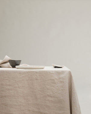 The Linen Tablecloth - Natural covers a table arranged with minimal ceramic dishes and bowls—some filled with dark contents—set against a plain, light background.