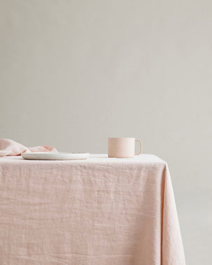 A pale pink mug and matching plate sit on a table covered with the Linen Tablecloth - Blush, set against a plain beige background.