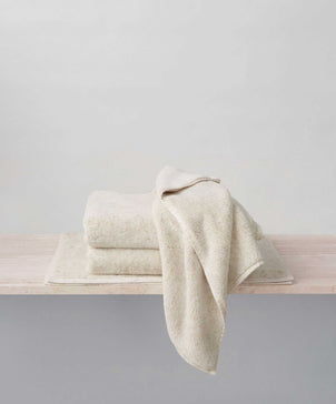 A stack of folded, off-white Woven Speckle Cotton Bath Sheet Bundles rests on a light wooden surface, with one towel elegantly draped over the stack against a plain, light background. Crafted from OEKO-TEX certified cotton, this quick-drying bundle provides both luxury and peace of mind.