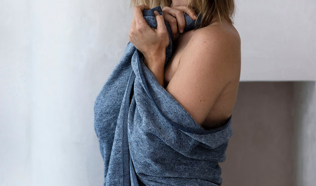 A female model as the Denim Bath Towel in Denim wrapped around herself, clasping it together at the front. She is standing sideways, facing away from the camera.