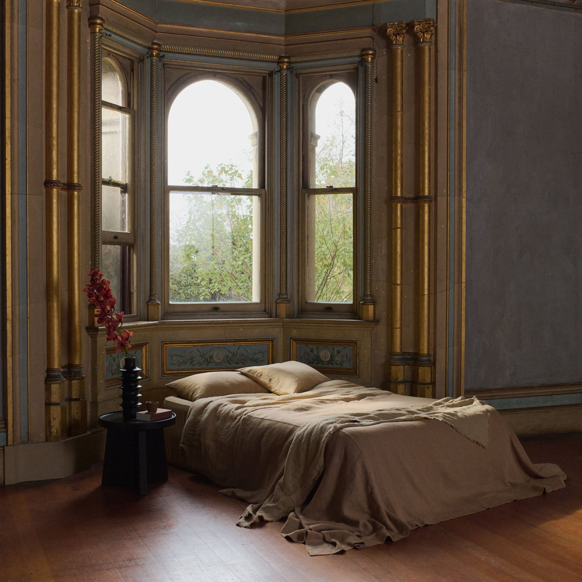 A bed with brown bedding is placed near tall arched windows in an ornate room with gold details. A small black table with a vase and red flowers sits next to the bed. Trees are visible through the windows.