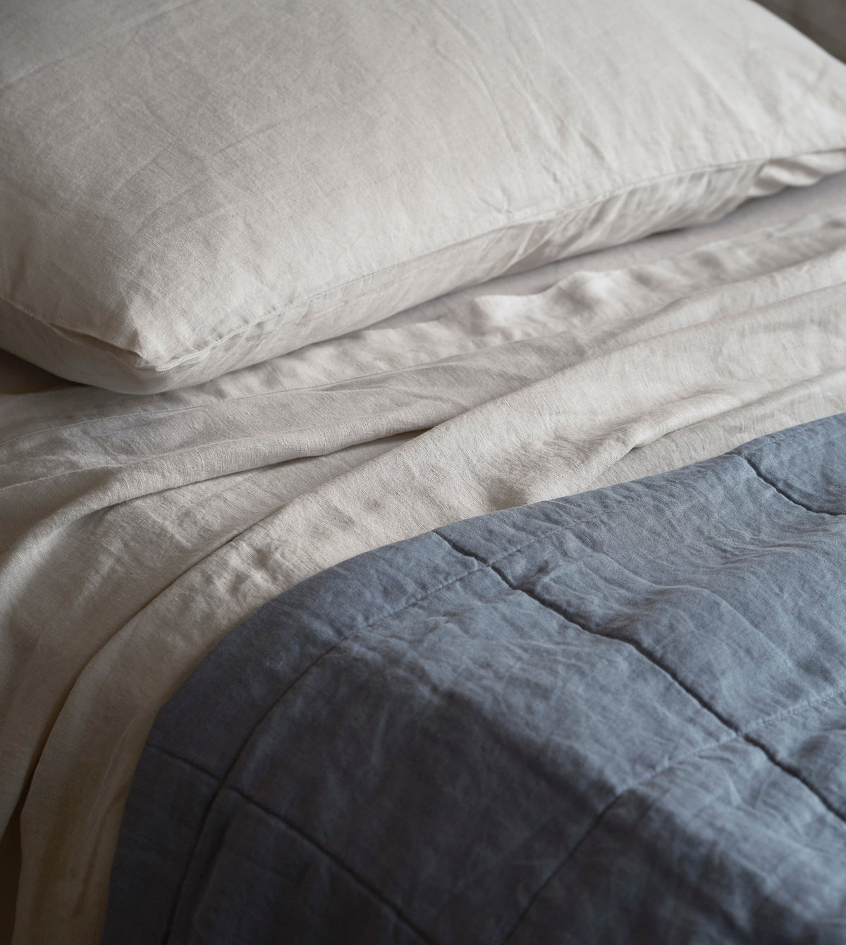 Close-up of a neatly made bed with a cream pillow and cream bedding, layered with a slate blue quilted blanket.