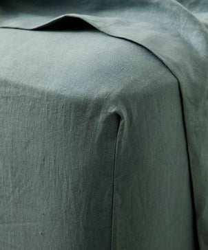 Close-up of the Linen Fitted Sheet - Bluestone, made from European flax, draped over a corner. The green linen’s natural texture and visible fold create a vintage look.