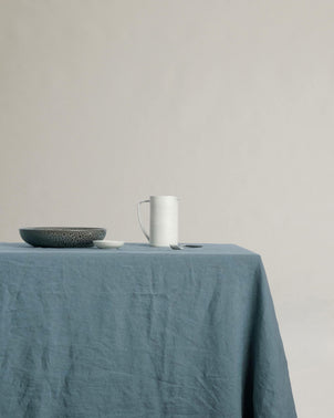 A table draped with the Linen Tablecloth - Bluestone holds a gray bowl, a small white dish, a fork, and a white pitcher, all set against a plain beige background.