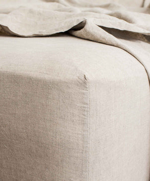 Close-up of a bed corner with the Linen Fitted Sheet - Natural made from European flax. The textured, soft beige fabric adds a vintage touch, with the fitted sheet neatly in place beneath a loosely draped flat sheet.