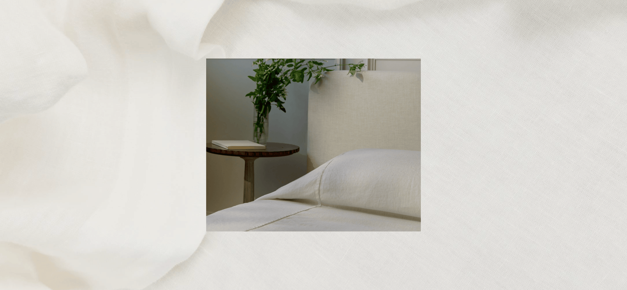 A neatly made bed with white linens sits next to a round wooden nightstand holding a closed book and a glass vase with green leafy branches.