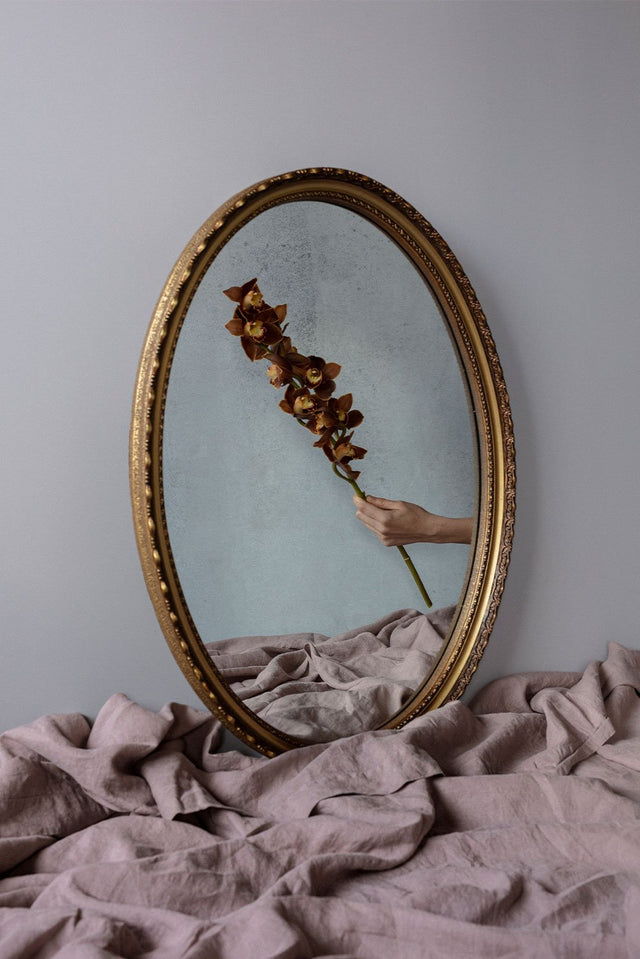 Image shows a mirror resting against a wall reflecting a hand holding a small branch with flowers and leaves