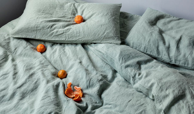 A bed styled with a Linen Duvet Cover in Sage and a Linen Fitted Sheet in Sage. Atop the bed are some mandarins, carefully placed. 