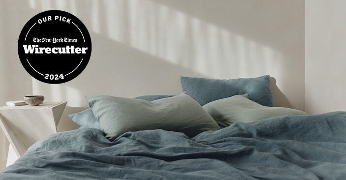 A neatly made bed with blue-green bedding and pillows sits in soft sunlight. Next to the bed is a small white table with a bowl. A Wirecutter Our Pick 2024 badge appears in the upper left corner.