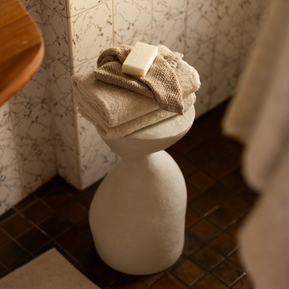 A stack of folded beige and cream towels sits on a sculptural white pedestal stool in a bathroom setting with patterned tile walls and dark brown floor tiles. A bar of soap rests on top of the towels.