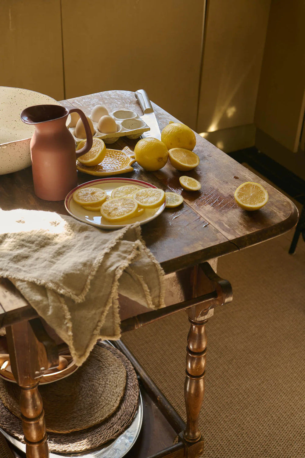 A rustic wooden table holds sliced lemons, a ceramic plate, eggs, a pink pitcher, a beige bowl, and a textured cloth, all bathed in warm, natural light.