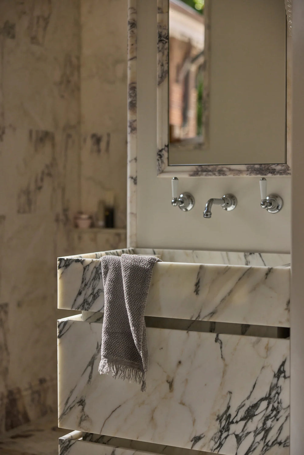 A marble bathroom vanity with three drawers, two faucet handles, and a gray towel draped over the edge. A mirror above the sink reflects part of a window and the marble wall.