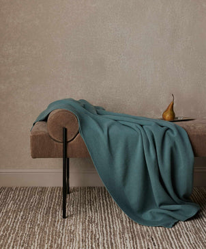 A 100% NZ Merino Wool Throw in Bluestone is draped over a brown upholstered bench with black metal legs. A single pear rests on a small plate at one end. The bench sits on a striped rug against a beige wall, highlighting Australian craftsmanship.