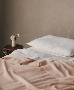 A neatly made bed with white pillows and the 100% NZ Merino Wool Throw - Blush. A small round side table holds a white vase with pink flowers, set against a plain beige wall that highlights Australian craftsmanship.