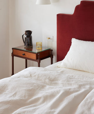 A neatly made bed with ivory linen pillowcases and a red upholstered headboard sits by a wooden nightstand with a ceramic pitcher and glass tumbler, creating a vintage feel against the white wall.
