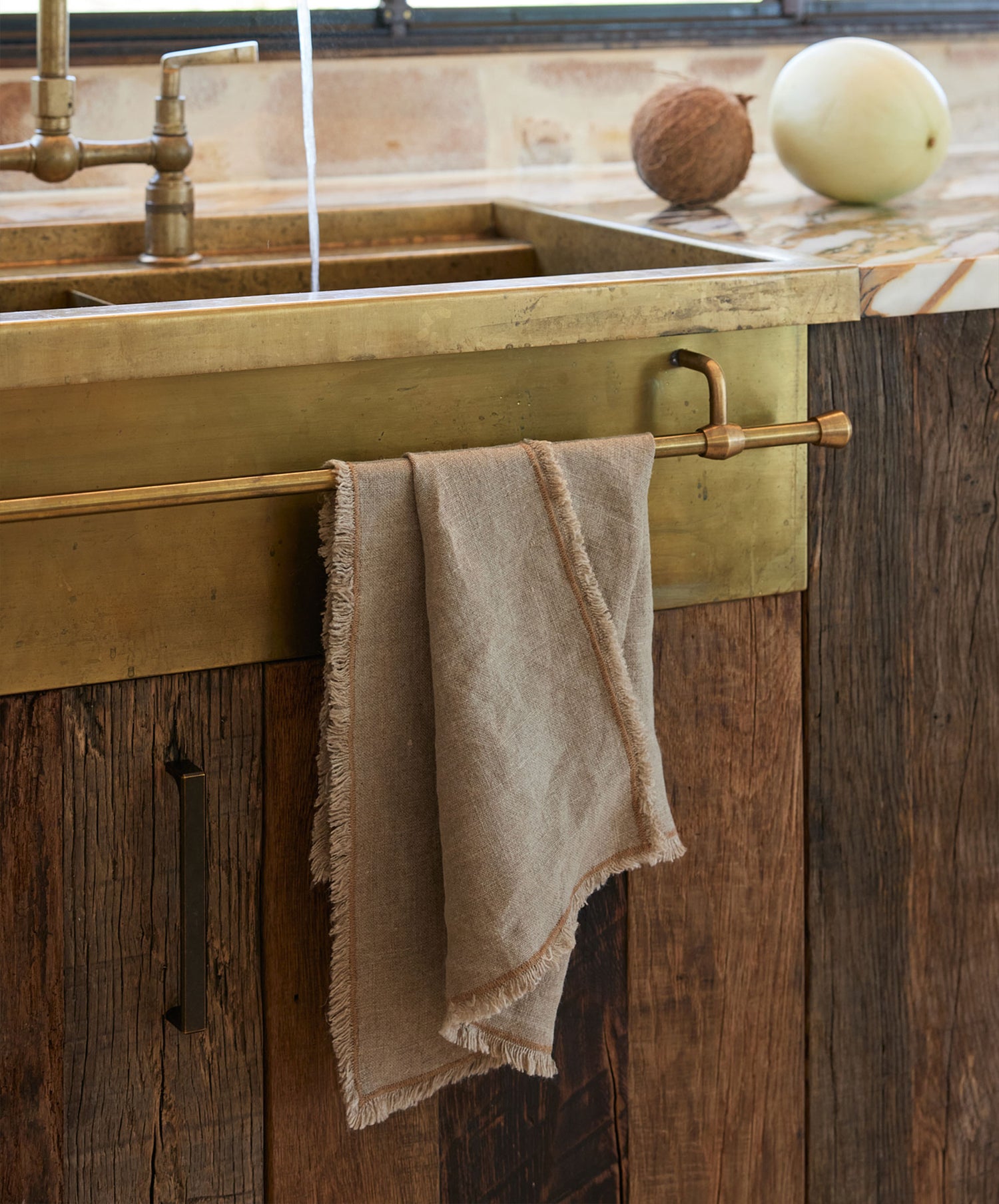 The Fia Kitchen Towel - Bronze, made of heavyweight linen in Portugal, hangs on a brass rod below a metal sink, with a melon and coconut resting on the rustic wooden countertop above.