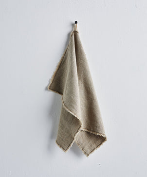 The Fia Kitchen Towel - Natural (20in x 28in) in beige linen with frayed edges hangs from a small hook on a plain white wall.