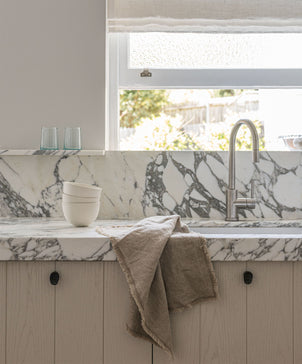 A marble kitchen countertop with a built-in sink, silver faucet, stacked white bowls, two glasses, and a Fia Kitchen Towel - Natural draped over the edge. Sunlight filters through the window above.