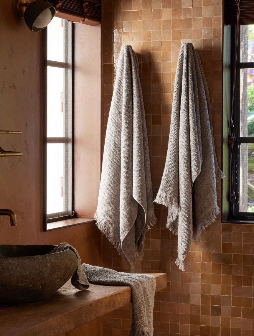 Two gray textured towels hang on hooks against a wall of brown tiles in a bathroom with a stone sink and sunlight coming through two windows. Another towel is draped over the counter.