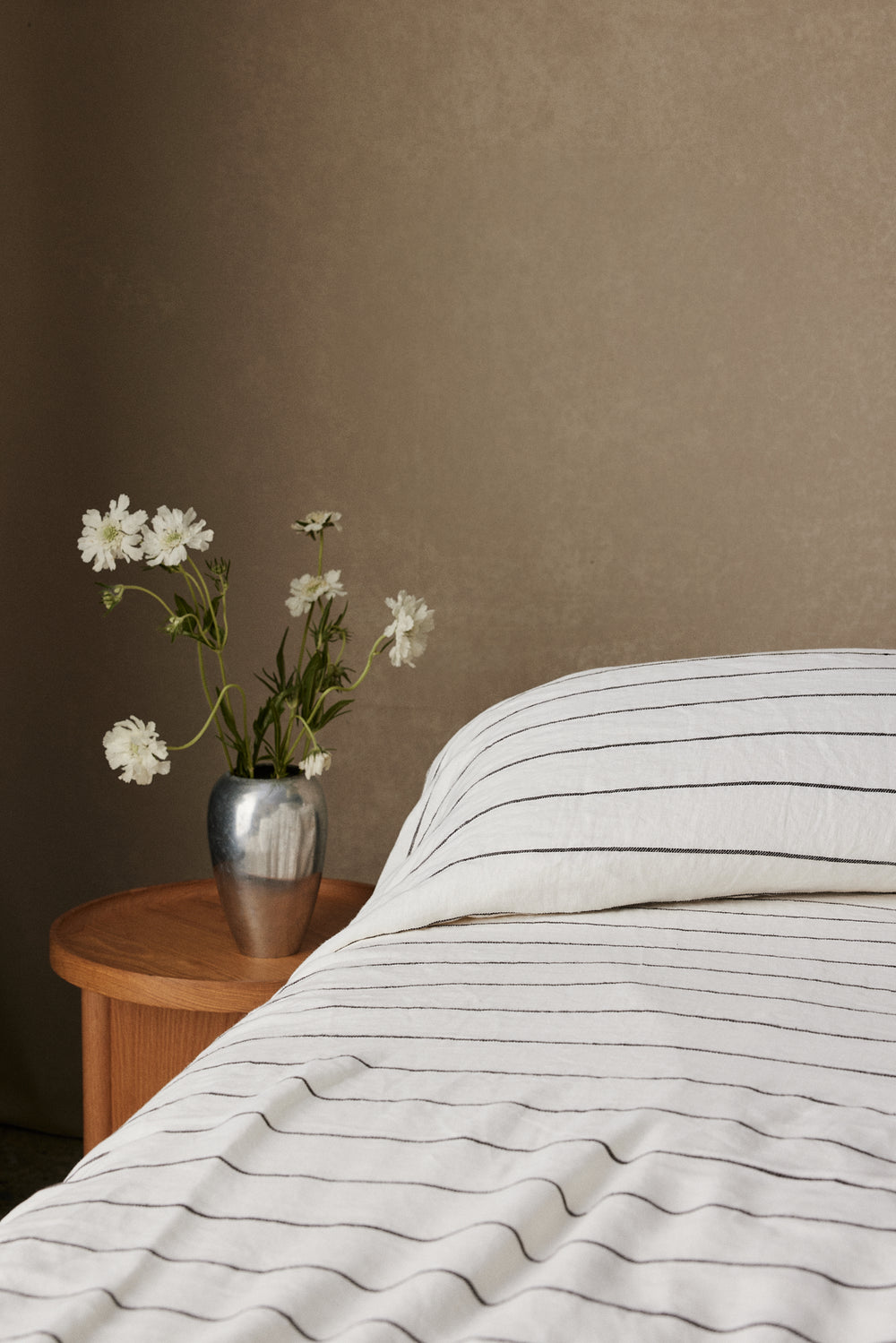 A bed with white and gray striped bedding is next to a small wooden nightstand holding a metallic vase with white flowers, set against a plain beige wall.