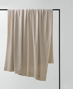 The 100% NZ Merino Wool Throw in Oatmeal, with a subtle texture, is draped over a sleek black metal frame on a plain white background, highlighting its premium quality and refined craftsmanship.