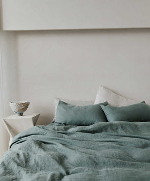A bed with sage green bedding and Set of 2 Linen Pillowcases in Bluestone rests by a light wall. A white geometric nightstand holds a textured bowl, creating a minimalist, serene room with subtle vintage charm.
