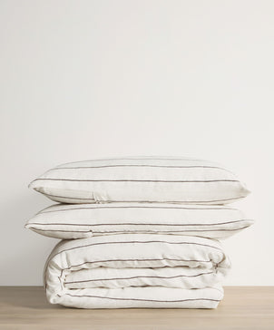 Two white pillows with thin black stripes rest on a folded Linen Duvet Cover Set - Truffle Stripe, all arranged on a light wooden surface with a plain white background.