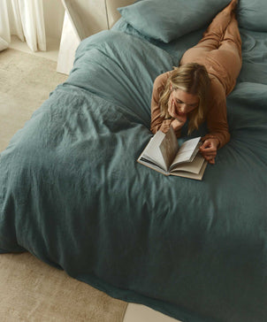 A woman relaxes on a bed with the Linen Fitted Sheet in Bluestone, made from European flax, reading a book in a softly lit, cozy bedroom with vintage charm.