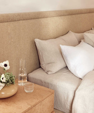 A cozy bed dressed in the Linen Fitted Sheet - Natural sits by a wooden nightstand with a glass of water, carafe, and beige vase of white flowers, creating a soft vintage ambiance.