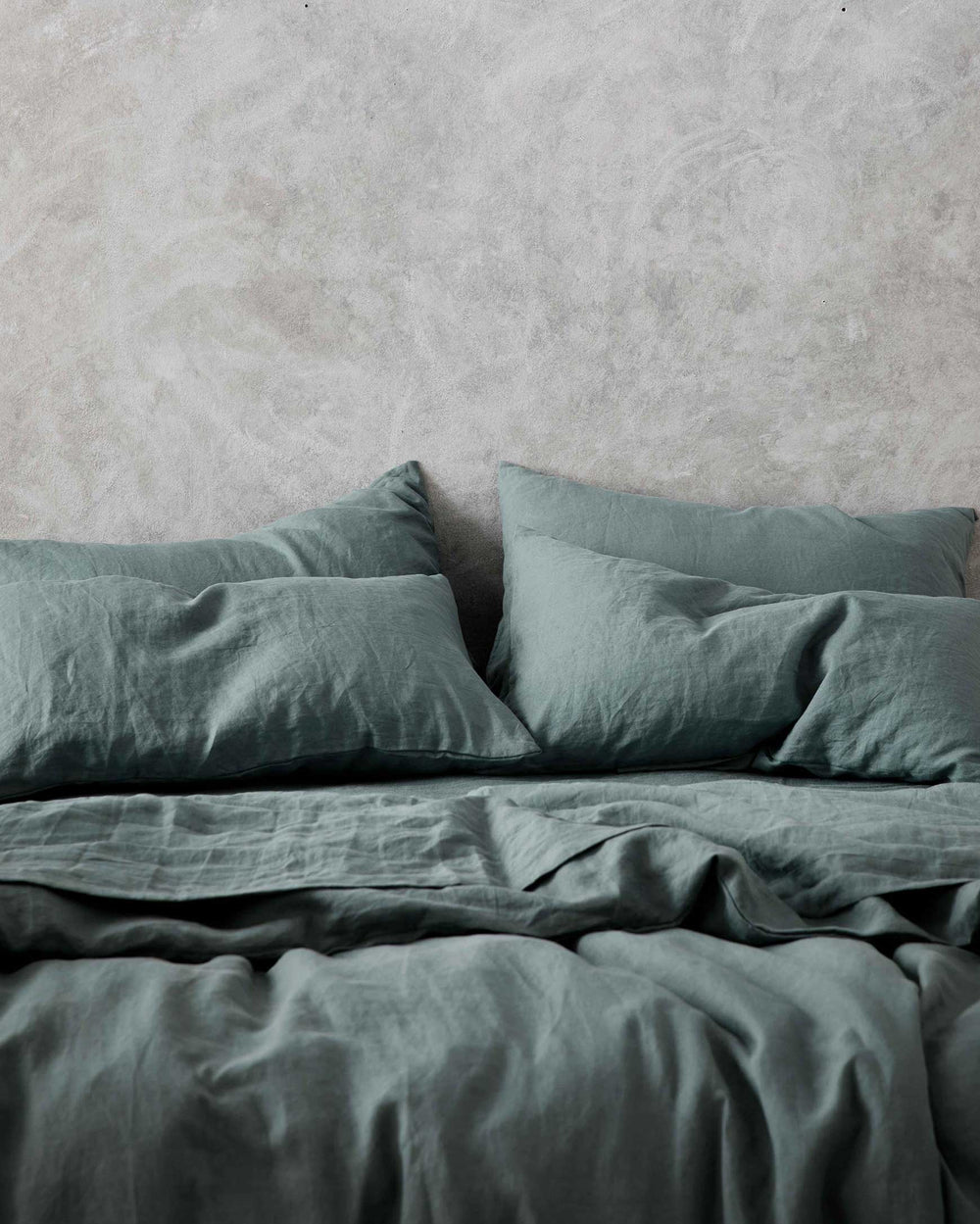 A neatly made bed with dark green bedding and two pillows, set against a textured light gray wall.