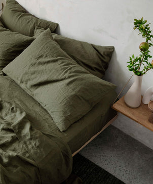 A neatly made bed with a Forest Linen Fitted Sheet sits by a small wooden nightstand topped with a white vase of green branches; the floor is gray concrete.