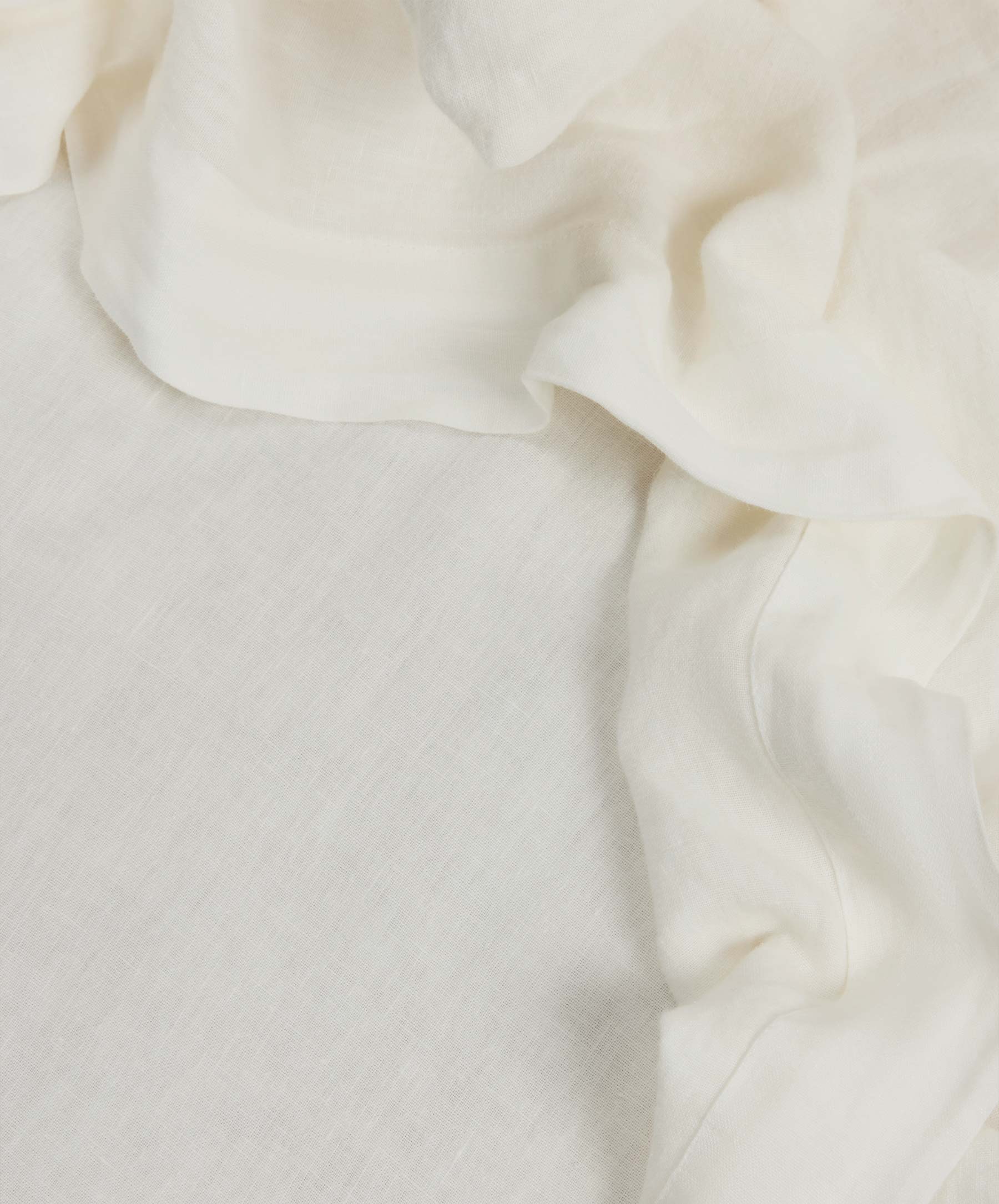 The Linen Fitted Sheet - Ivory, crafted from soft European flax linen in an off-white hue, drapes gracefully atop a matching surface, showcasing gentle folds and creases for a minimal and elegant look that is naturally temperature regulating.