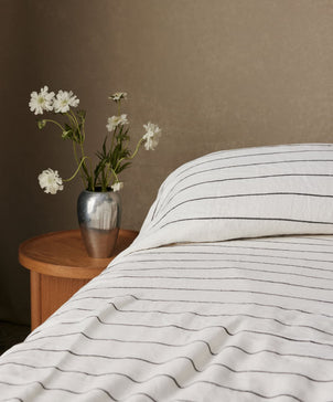 Striped bedding on a bed with a vase of flowers on a nightstand against a brown wall.