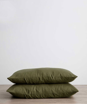 A set of 2 linen pillowcases in Forest, crafted from European flax, is stacked on a wooden floor against a white wall, adding a vintage touch to any room.