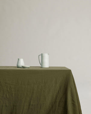 A Linen Tablecloth - Forest drapes over the table, displaying a pale ceramic pitcher, a small cup, and a shallow dish on its surface against a simple, light-colored background.