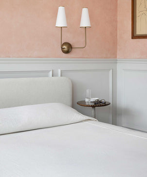 A neatly made bed with a light-colored headboard features the Heavyweight Linen Bedcover - Ivory. Beside it is a small round table with water, books, and glasses. The wall is pink above white paneling, with a modern sconce above the table.