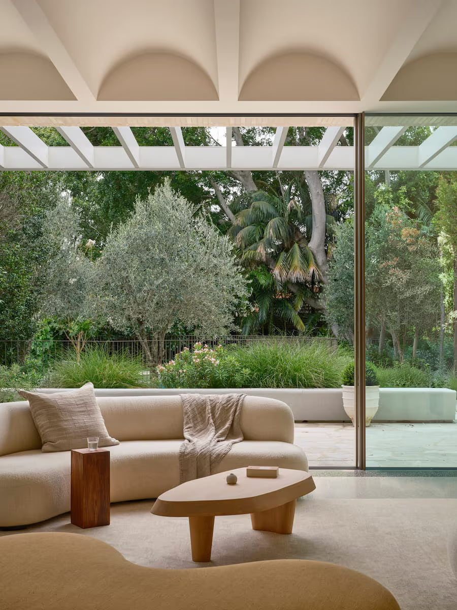 A modern living room with a curved beige sofa, wooden table, and blanket, large windows open to a lush green garden with trees and plants outside. Natural light fills the room, creating a calm, inviting atmosphere.