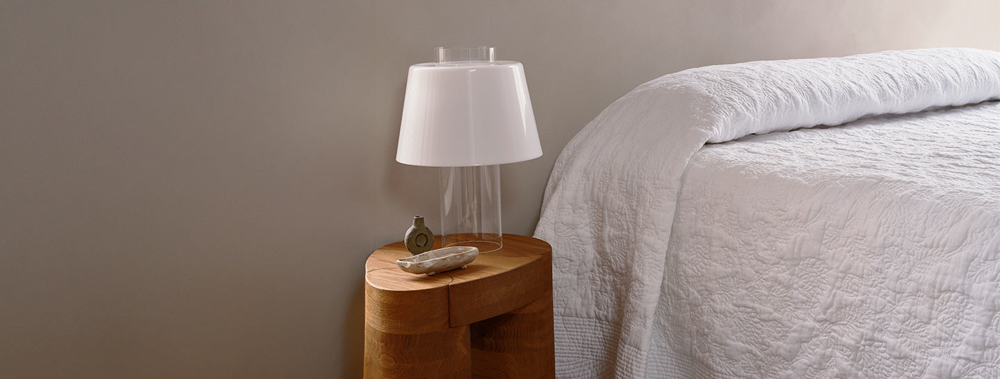 A wooden bedside table with a modern white lamp, a small dish, and a perfume bottle next to a neatly made bed with a white quilted cover. The background wall is plain and light-colored.