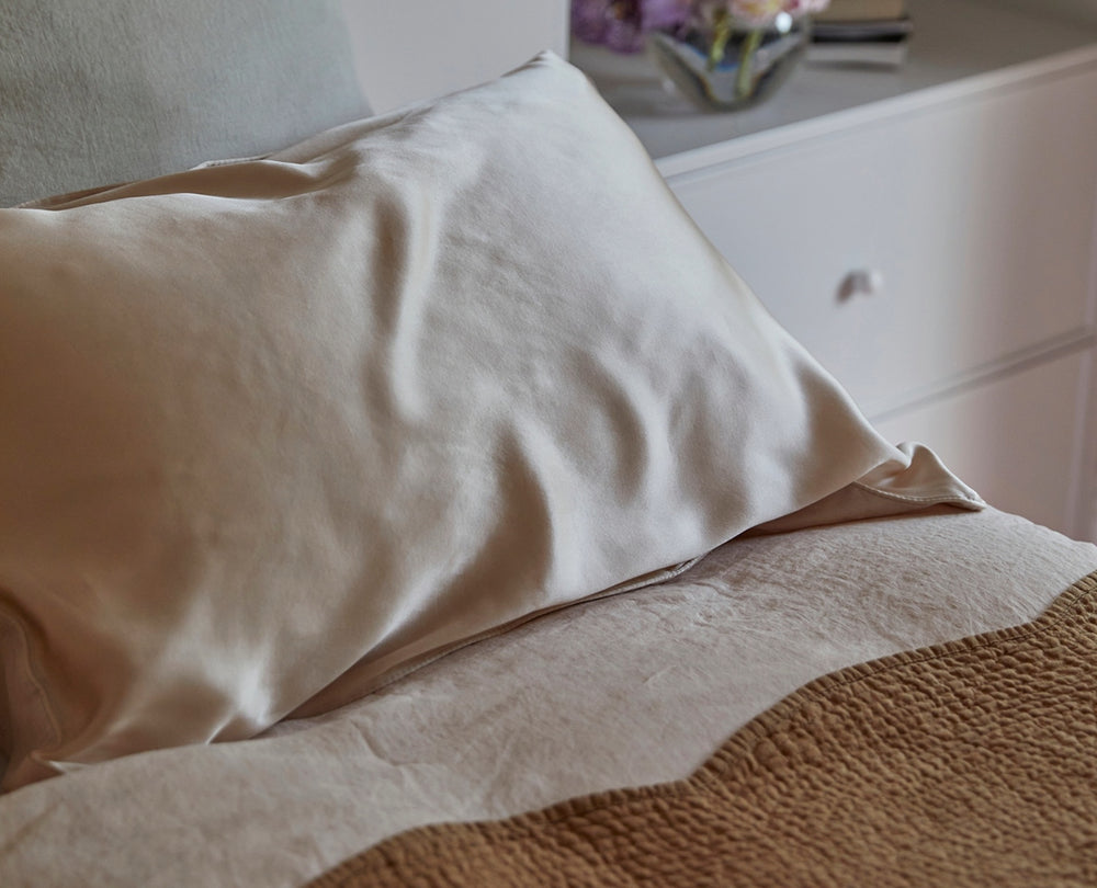 A close-up of a bed with a cream-colored satin pillow on light bedding. A beige textured blanket is partially visible, and a white nightstand with a glass vase of flowers is in the background.