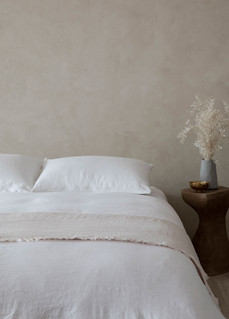 A neatly made bed with white sheets and pillows stands next to a wooden side table holding a white vase with dried flowers and a small golden bowl, set against a light beige wall.