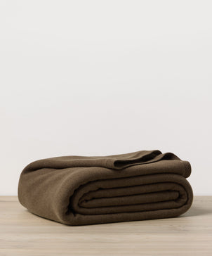 A 100% NZ Merino Wool Blanket - Truffle, neatly folded, rests on a light wooden surface with a plain white background.