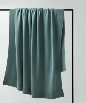 A 100% NZ Merino Wool Throw in Bluestone hangs over a simple black metal rod against a white background. The smooth fabric, expertly crafted in New Zealand, drapes with gentle folds for a relaxed, cozy look.